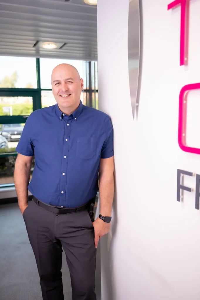 Colin Tapscott, Everyday Leader coach, standing portrait beside a branded office wall