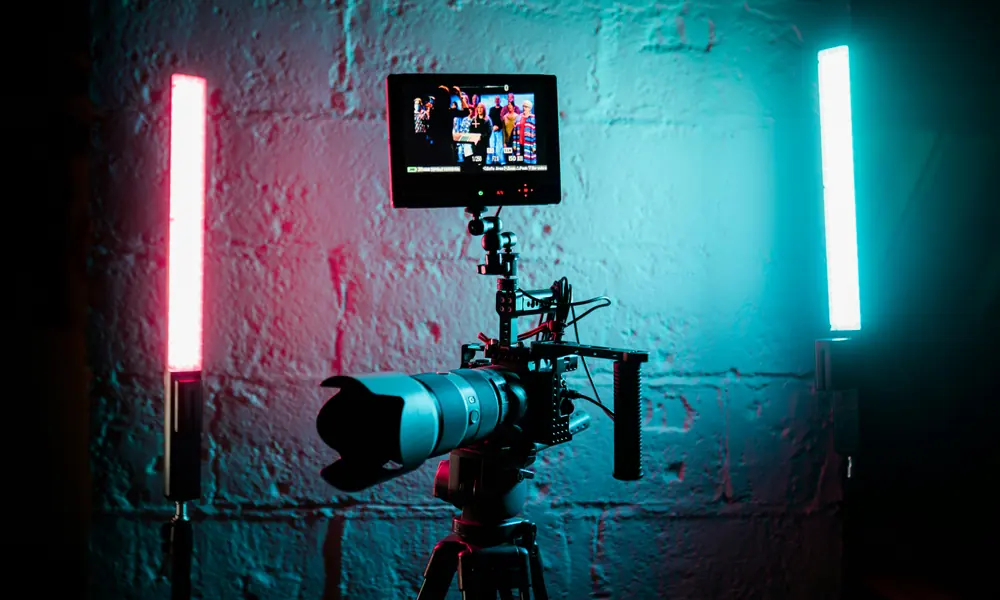 Cinema camera on a tripod between magenta and cyan studio tube lights against a white brick wall on a video production set